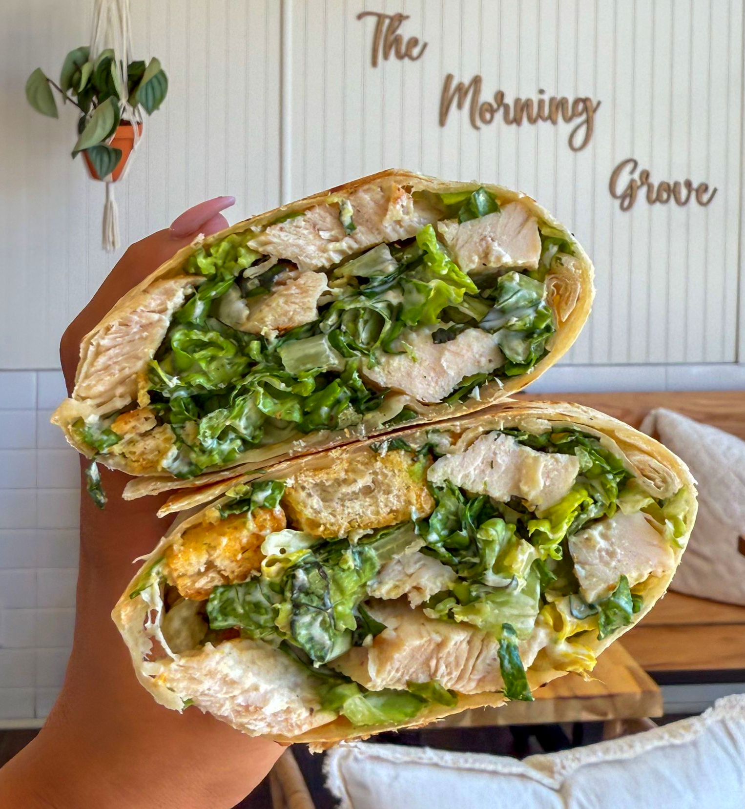 Chicken Caesar wrap, cut in half, held in hand. Romaine, croutons, and chicken visible. Setting is 